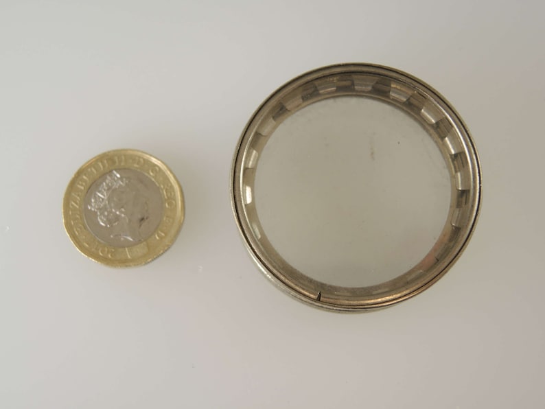 Puede incluir: Un recipiente redondo de metal plateado con una tapa de vidrio transparente. El recipiente es aproximadamente del tama&ntilde;o de una moneda de un euro.