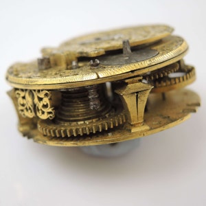 May include: Close-up of a gold-toned pocket watch mechanism with intricate gears and a decorative design. The watch is disassembled, revealing the inner workings.