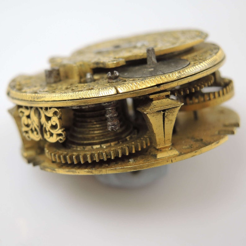 May include: Close-up of a gold-toned pocket watch mechanism with intricate gears and a decorative design. The watch is disassembled, revealing the inner workings.