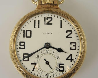 May include: A gold-tone pocket watch with a white face, black Arabic numerals, and black hands. The dial features the text ELGIN and a smaller sub-dial for seconds at the bottom. The watch has a textured gold-tone bezel and a top winding crown.