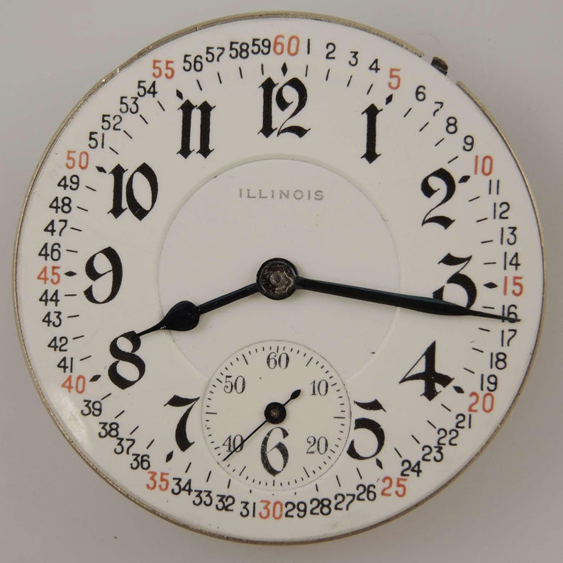 May include: A vintage Illinois pocket watch face with a white background, black numerals, and a smaller seconds dial. The watch face has black hands and a metal casing. The numbers 15, 30, 45, and 60 are in red.