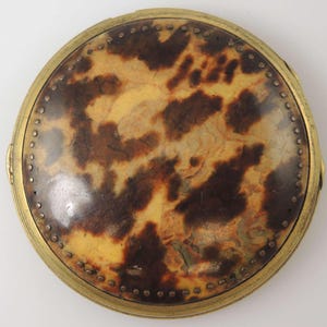 Puede incluir: Un compacto redondo con estampado de caparazón de tortuga, con un borde de latón y un borde decorativo de pequeños remaches de latón espaciados uniformemente.