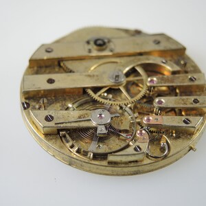May include: Detailed view of a vintage watch movement, revealing intricate gears, springs, and levers. The brass components are detailed with small red jewels and screws. The watch mechanism is set against a white background.