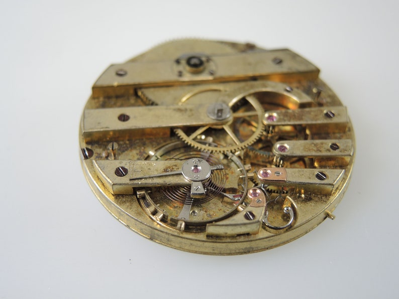 May include: Detailed view of a vintage watch movement, revealing intricate gears, springs, and levers. The brass components are detailed with small red jewels and screws. The watch mechanism is set against a white background.