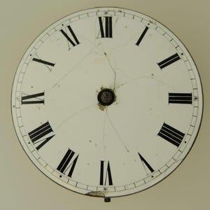 Puede incluir: Una esfera de reloj vintage con n&uacute;meros romanos. La esfera blanca tiene n&uacute;meros negros y marcadores de minutos. La esfera est&aacute; agrietada, mostrando signos de edad y desgaste. Una peque&ntilde;a pieza de metal est&aacute; en la parte inferior.