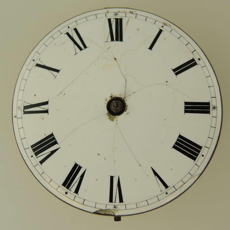 Puede incluir: Una esfera de reloj vintage con n&uacute;meros romanos. La esfera blanca tiene n&uacute;meros negros y marcadores de minutos. La esfera est&aacute; agrietada, mostrando signos de edad y desgaste. Una peque&ntilde;a pieza de metal est&aacute; en la parte inferior.