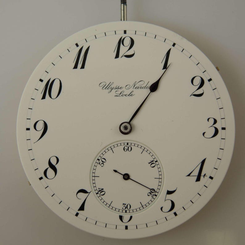 May include: A white pocket watch face with black Roman numeral hour markers and black hands. The watch face also features a smaller, white sub-dial with black markings. The watch face is inscribed with "Ulysse Nardin Locle".