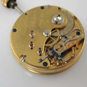 Puede incluir: Vista cercana del funcionamiento interno de un reloj de bolsillo de oro. La esfera del reloj es visible, con engranajes intrincados y un mecanismo de cuerda.