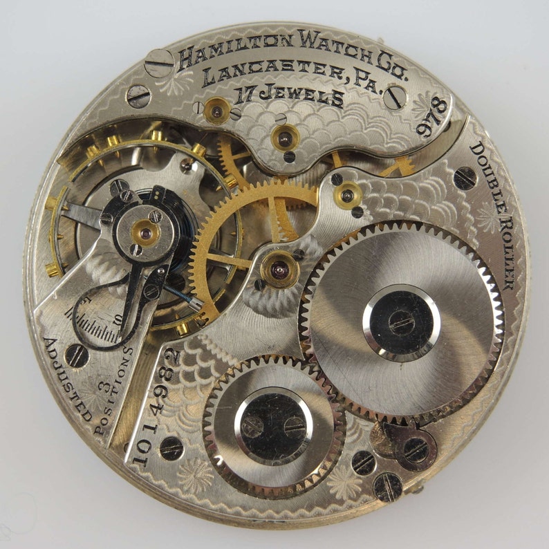 May include: Close-up of a silver pocket watch movement with gold gears. The watch face is engraved with "Hamilton Watch Co. Lancaster, Pa. 17 Jewels" and "Double Roller". The watch is numbered "1014982" and "978".