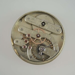 May include: Close-up view of the inner workings of a silver pocket watch. The watch face is removed, revealing the intricate gears and mechanisms. The watch has a silver finish and features a variety of gears and springs.