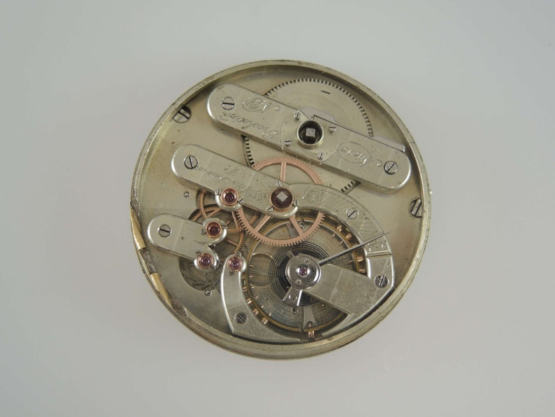 May include: Close-up view of the inner workings of a silver pocket watch. The watch face is removed, revealing the intricate gears and mechanisms. The watch has a silver finish and features a variety of gears and springs.