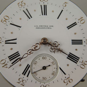 May include: A white pocket watch face with black Roman numeral hour markers and black minute markers. The watch face has a gold-colored inscription that reads "C.R. SMITHE SON, PHILADELPHIA."