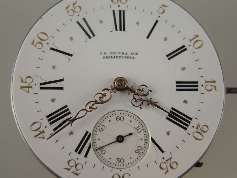 May include: A white pocket watch face with black Roman numeral hour markers and black minute markers. The watch face has a gold-colored inscription that reads "C.R. SMITHE SON, PHILADELPHIA."