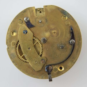 May include: Close-up of a vintage watch movement. The brass-colored mechanism features gears, screws, and a balance wheel. The design includes a curved, dark-colored spring and a small, silver-colored gear.