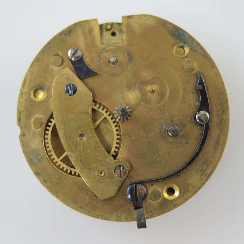 May include: Close-up of a vintage watch movement. The brass-colored mechanism features gears, screws, and a balance wheel. The design includes a curved, dark-colored spring and a small, silver-colored gear.