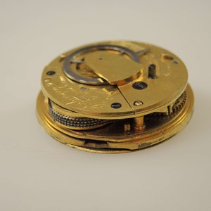 May include: A gold-colored pocket watch movement with intricate gears and a winding mechanism. The watch is open and the inner workings are visible.