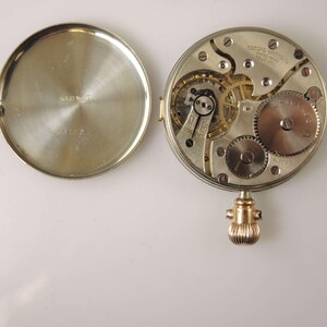 Puede incluir: Reloj de bolsillo abierto que muestra su mecanismo interno. La tapa trasera plateada est&aacute; abierta, revelando engranajes, resortes y otros componentes. La tapa trasera tiene la inscripci&oacute;n "SOLID NICKEL". La corona es dorada.