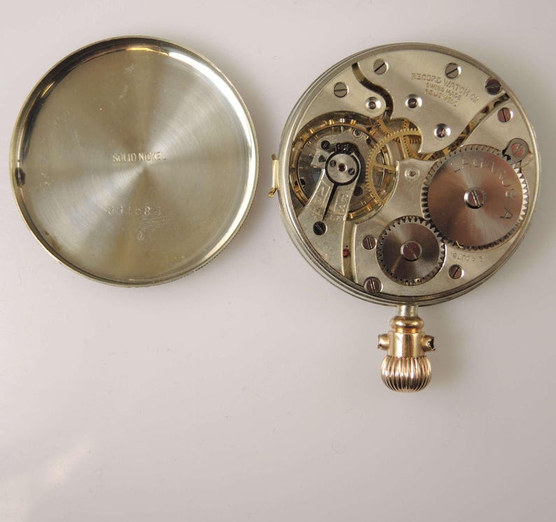Puede incluir: Reloj de bolsillo abierto que muestra su mecanismo interno. La tapa trasera plateada est&aacute; abierta, revelando engranajes, resortes y otros componentes. La tapa trasera tiene la inscripci&oacute;n "SOLID NICKEL". La corona es dorada.