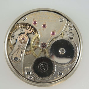 K&ouml;nnte beinhalten: Nahaufnahme des Innenlebens einer silbernen Taschenuhr mit der Aufschrift "Laforge & Valentine" und "Swiss" auf dem Zifferblatt. Die Uhr hat eine Reihe von Zahnr&auml;dern und einen schwarzen Metall-Aufzugsschl&uuml;ssel.