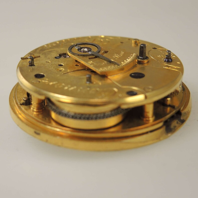 Puede incluir: Un movimiento de reloj de bolsillo dorado con engranajes intrincados y un mecanismo de cuerda. La esfera del reloj es visible, pero las manecillas faltan.