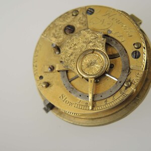 May include: Close-up of a gold-toned pocket watch movement with intricate details. The watch face features the inscription "J. Howells 1763" and a scale labeled "Slow" with numbers 1 through 6.