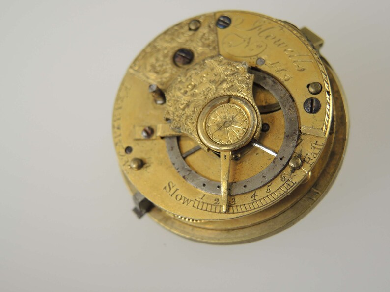 May include: Close-up of a gold-toned pocket watch movement with intricate details. The watch face features the inscription "J. Howells 1763" and a scale labeled "Slow" with numbers 1 through 6.