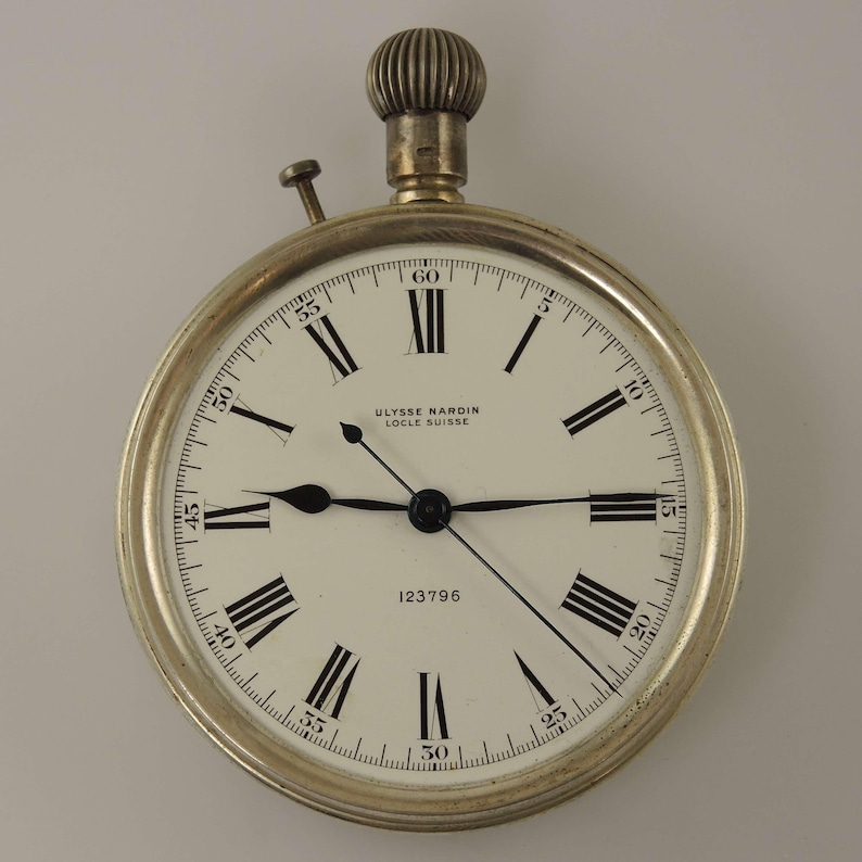 K&ouml;nnte beinhalten: Eine silberfarbene Taschenuhr mit wei&szlig;em Zifferblatt und schwarzen r&ouml;mischen Ziffern. Die Uhr tr&auml;gt den Text "ULYSSE NARDIN LOCLE SUISSE" und die Nummer "123796". Sie hat eine Krone und einen kleinen Knopf. Durchmesser ca. 5 cm.