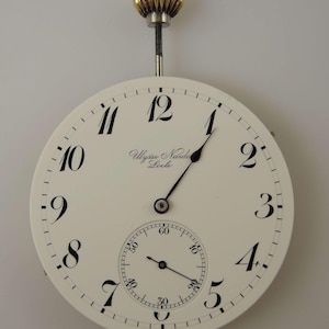 May include: A white pocket watch with black Roman numeral hour markers and black hands. The watch face also includes a smaller subdial with black markings. The watch is labeled "Ulysse Nardin Locle".