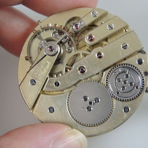 Puede incluir: Primer plano de un movimiento de reloj de bolsillo de tono dorado con engranajes intrincados y una palanca. El movimiento se sostiene en una mano y la esfera del reloj no es visible.