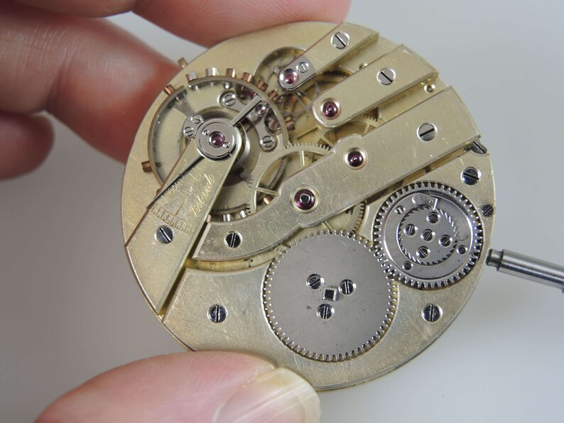 Puede incluir: Primer plano de un movimiento de reloj de bolsillo de tono dorado con engranajes intrincados y una palanca. El movimiento se sostiene en una mano y la esfera del reloj no es visible.