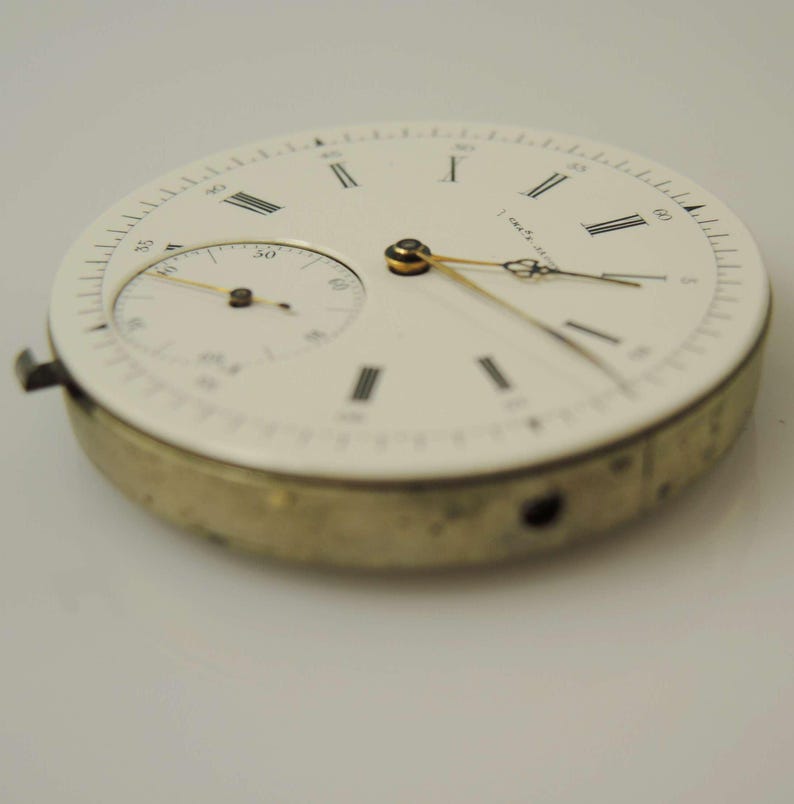 May include: A vintage watch face with a white dial and black Roman numerals. The watch face has a smaller subdial and gold-coloured hands. The outer casing is a gold colour. The watch face is a classic timepiece component.