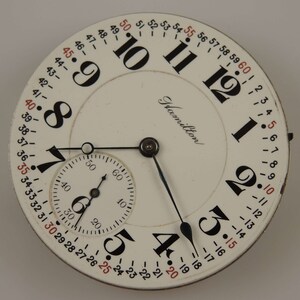 May include: A white pocket watch with black Roman numeral hour markers and black hands. The watch face also has a smaller, concentric circle with black Arabic numeral markers and a black hand. The watch face is labeled "Hamilton".
