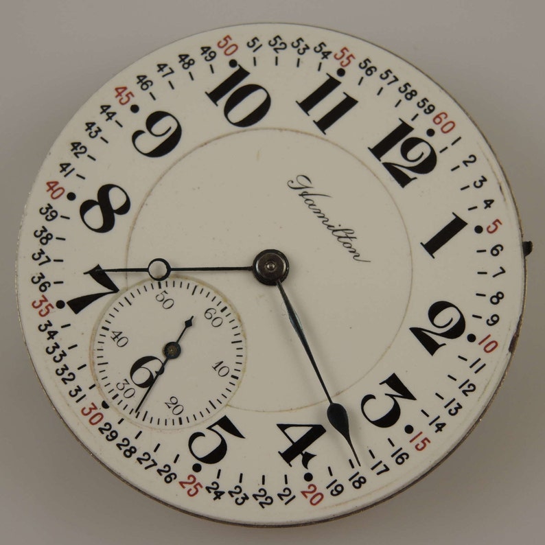 May include: A white pocket watch with black Roman numeral hour markers and black hands. The watch face also has a smaller, concentric circle with black Arabic numeral markers and a black hand. The watch face is labeled "Hamilton".