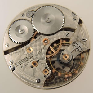 May include: Close-up of a vintage watch movement with intricate gears and components. The silver-toned mechanism features the text "A.W.C. Waltham, Mass." and "17 Jewels Adjusted."