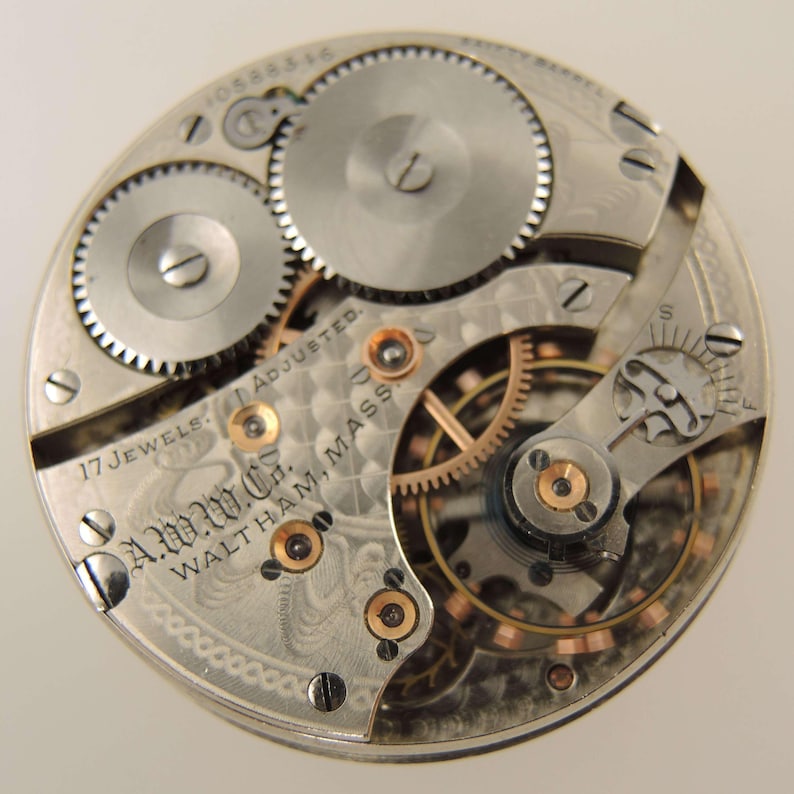 May include: Close-up of a vintage watch movement with intricate gears and components. The silver-toned mechanism features the text "A.W.C. Waltham, Mass." and "17 Jewels Adjusted."