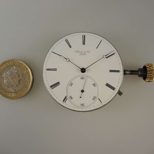 May include: A white pocket watch face with black Roman numeral hour markers and black hands. The watch face has a smaller sub-dial with black markings. The watch face is surrounded by a silver-colored metal case. The watch has a gold-colored winding key.