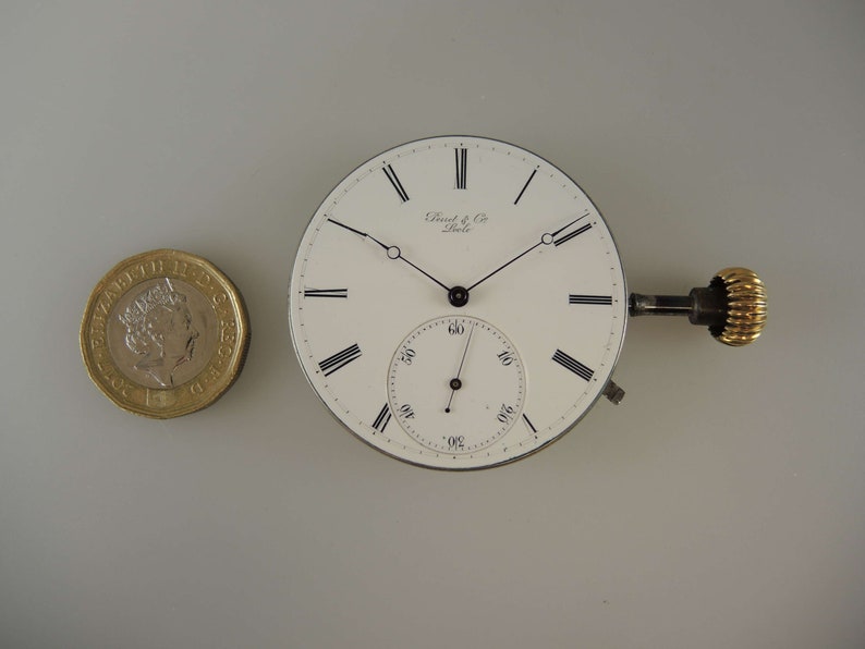 May include: A white pocket watch face with black Roman numeral hour markers and black hands. The watch face has a smaller sub-dial with black markings. The watch face is surrounded by a silver-colored metal case. The watch has a gold-colored winding key.