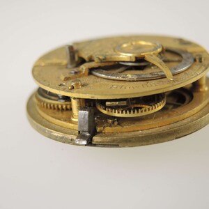 May include: Close-up of a gold-toned pocket watch movement with intricate gears and a silver-toned plate. The watch is disassembled and the inner workings are visible.