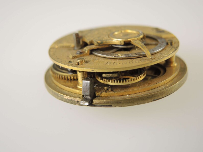 May include: Close-up of a gold-toned pocket watch movement with intricate gears and a silver-toned plate. The watch is disassembled and the inner workings are visible.