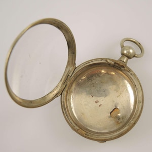 Könnte beinhalten: Silberne Taschenuhr mit einem aufklappbaren Deckel, der das Innere der Uhr freilegt. Das Zifferblatt der Uhr fehlt. Die Uhr hat einen kleinen, runden, weißen Knopf im Inneren des Gehäuses.