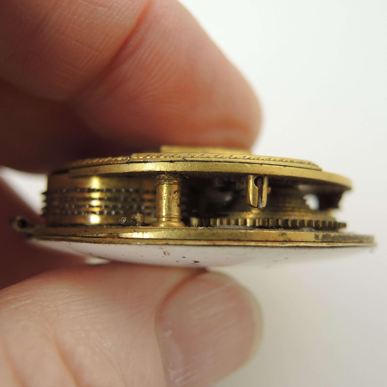 Puede incluir: Primer plano del mecanismo de un reloj de bolsillo antiguo. El reloj presenta intrincados engranajes y componentes de color dorado. El reloj tiene una esfera blanca.