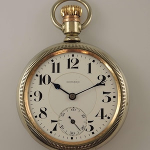 Könnte beinhalten: Eine silberne Taschenuhr mit weißem Zifferblatt, schwarzen Ziffern und blauen Zeigern. Die Uhr hat eine goldfarbene Lünette und Krone. Das Wort "HOWARD" ist auf dem Zifferblatt aufgedruckt. Ein kleineres Zifferblatt befindet sich unten.