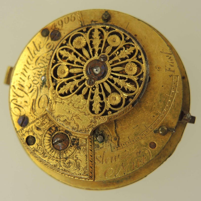 Puede incluir: Primer plano de un movimiento de reloj de oro antiguo. El intrincado mecanismo presenta dise&ntilde;os florales ornamentados, texto grabado y una joya central. La esfera del reloj muestra el texto "P. Grimalde London" y "4906".