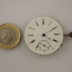 May include: A white watch face with black Roman numerals and a smaller seconds dial. The watch face has a gold-colored winding stem and is next to a gold-colored coin.