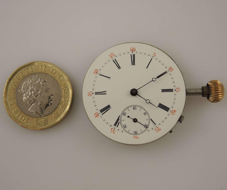 May include: A white watch face with black Roman numerals and a smaller seconds dial. The watch face has a gold-colored winding stem and is next to a gold-colored coin.