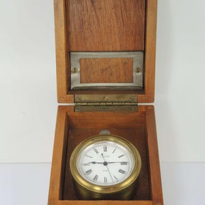 May include: A brass clock with a white face and Roman numerals, inside an open wooden box. The clock has a gold-coloured bezel and is set against the wood grain of the box. The box has a metal plate on the inside of the lid.