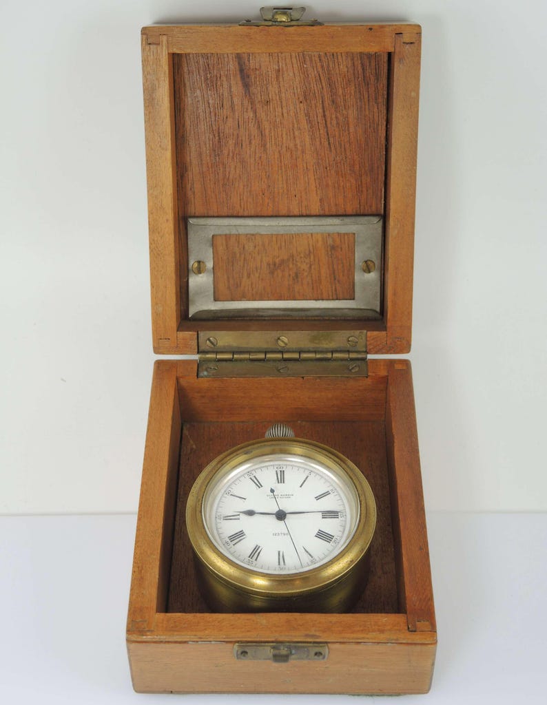 K&ouml;nnte beinhalten: Eine Vintage-Messing-Uhr mit wei&szlig;em Zifferblatt und r&ouml;mischen Ziffern, untergebracht in einer offenen Holzkiste. Die Uhr hat eine goldfarbene L&uuml;nette und ist vor der Holzmaserung der Kiste platziert. Die Kiste hat eine Metallplatte auf der Innenseite des Deckels.