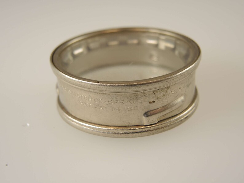 Puede incluir: Un anillo de caja de reloj de metal plateado con la inscripci&oacute;n "The Watch Co. of  S.F.  K.G.  U.S.A. Pat. May 14, 1902".