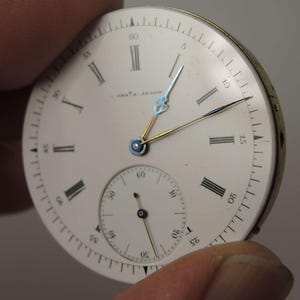 May include: A vintage watch face with a white background, black Roman numerals, and a smaller seconds dial. The watch hands are gold and blue. The text "CHA'S. JACOB" is printed on the face.