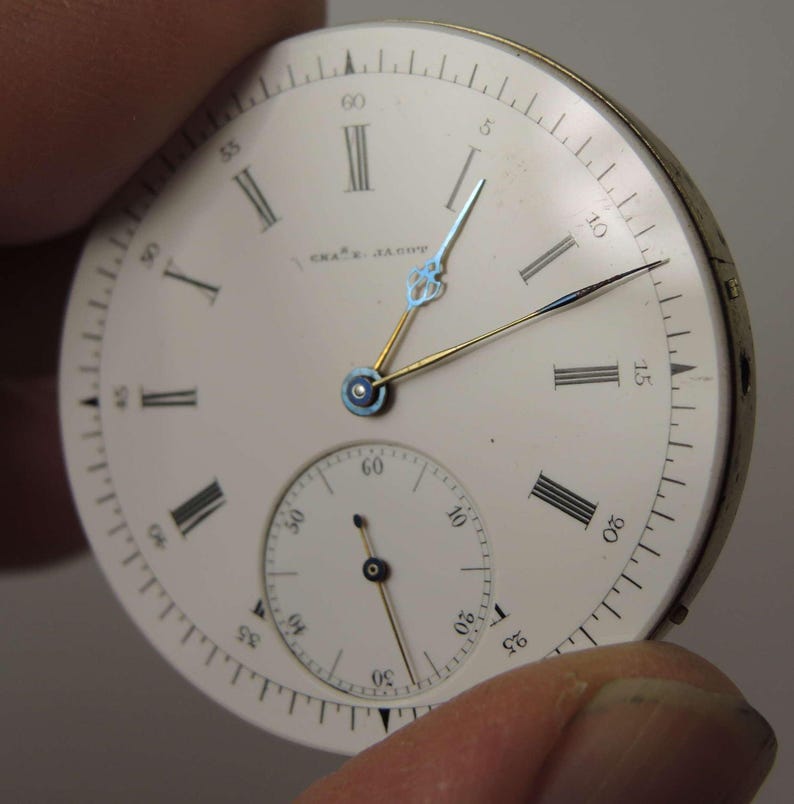 May include: A vintage watch face with a white background, black Roman numerals, and a smaller seconds dial. The watch hands are gold and blue. The text "CHA'S. JACOB" is printed on the face.